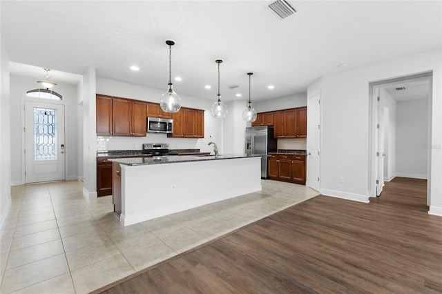 kitchen featuring appliances with stainless steel finishes, light wood-type flooring, backsplash, a kitchen island with sink, and hanging light fixtures