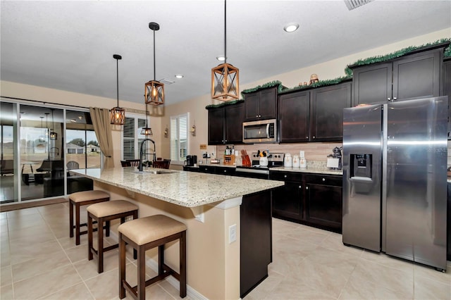 kitchen featuring stainless steel appliances, sink, light stone countertops, a center island with sink, and pendant lighting