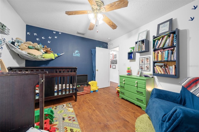 bedroom featuring a textured ceiling, ceiling fan, wood-type flooring, and a crib