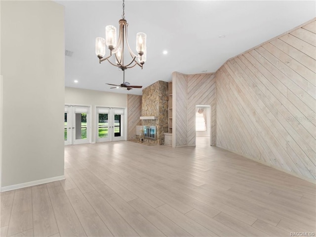 unfurnished living room with ceiling fan with notable chandelier, wood walls, a stone fireplace, and light wood-type flooring