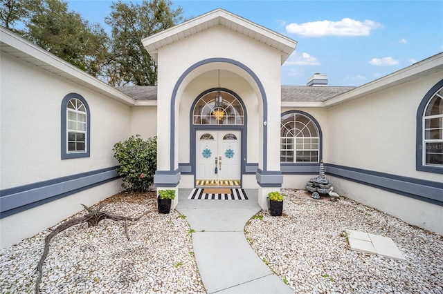 view of exterior entry with roof with shingles, a chimney, and stucco siding
