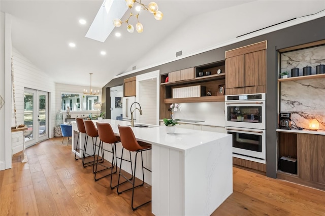 kitchen with vaulted ceiling with skylight, multiple ovens, a kitchen island with sink, pendant lighting, and an inviting chandelier