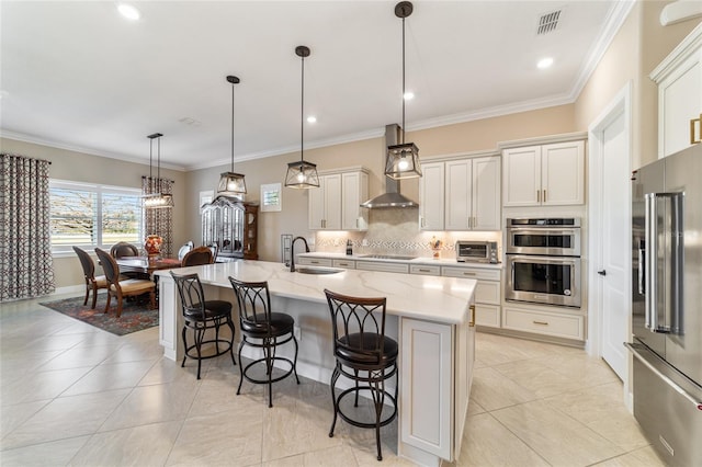 kitchen with sink, stainless steel appliances, wall chimney range hood, decorative light fixtures, and a kitchen island with sink