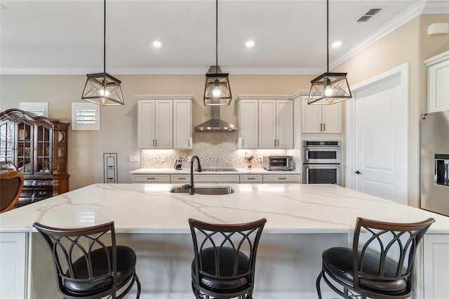 kitchen featuring wall chimney exhaust hood, white cabinetry, an island with sink, and hanging light fixtures