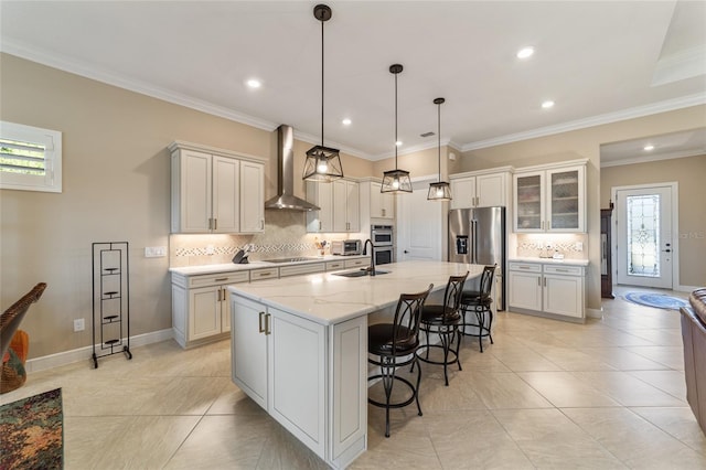 kitchen with appliances with stainless steel finishes, light stone counters, wall chimney exhaust hood, decorative light fixtures, and an island with sink