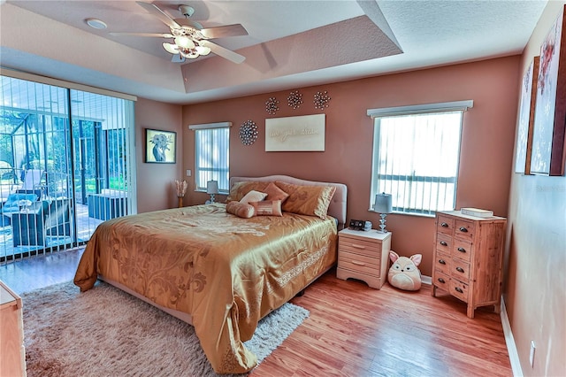 bedroom with ceiling fan, light hardwood / wood-style floors, a textured ceiling, access to outside, and a raised ceiling