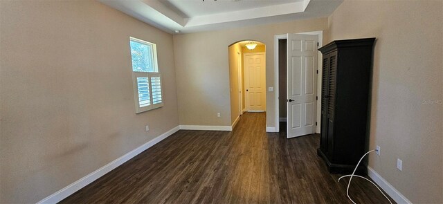 unfurnished bedroom with arched walkways, a raised ceiling, baseboards, and dark wood-style flooring
