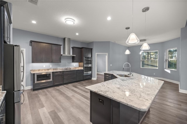 kitchen with pendant lighting, a large island with sink, sink, wall chimney exhaust hood, and appliances with stainless steel finishes