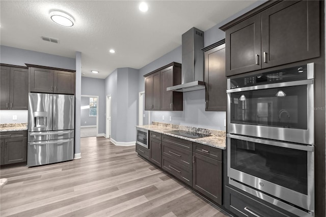 kitchen with light stone countertops, wall chimney range hood, a textured ceiling, dark brown cabinets, and appliances with stainless steel finishes