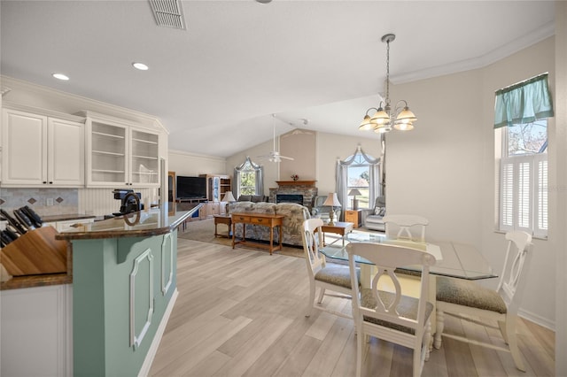 dining space featuring light wood-type flooring, a stone fireplace, vaulted ceiling, and crown molding