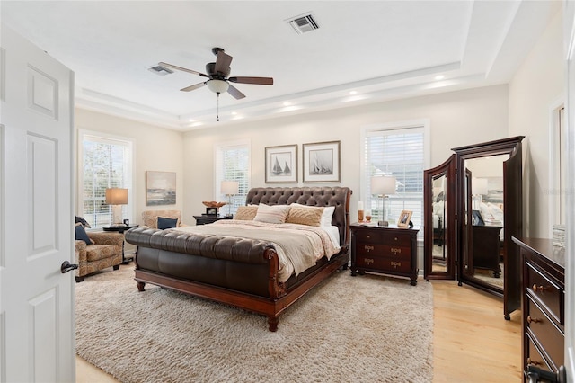 bedroom featuring ceiling fan, a tray ceiling, multiple windows, and light wood-type flooring