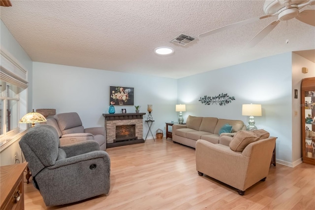 living room featuring ceiling fan, a textured ceiling, light hardwood / wood-style flooring, and a stone fireplace