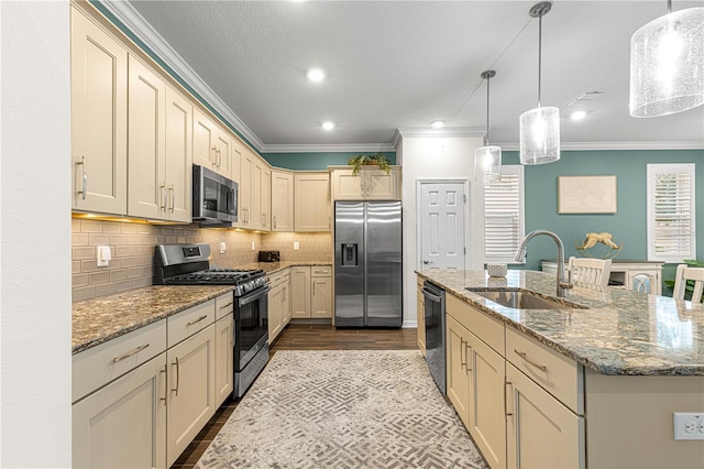 kitchen with an island with sink, appliances with stainless steel finishes, ornamental molding, pendant lighting, and sink