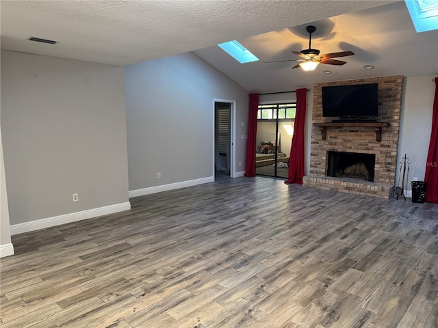 unfurnished living room featuring ceiling fan, hardwood / wood-style floors, vaulted ceiling with skylight, a textured ceiling, and a brick fireplace