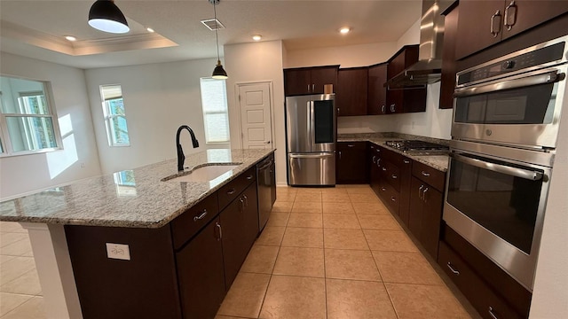 kitchen with sink, a center island with sink, a tray ceiling, stainless steel appliances, and wall chimney range hood