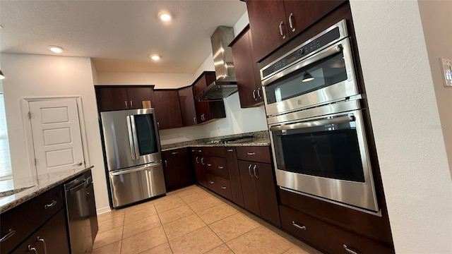 kitchen featuring light tile patterned floors, stainless steel appliances, light stone countertops, dark brown cabinets, and wall chimney exhaust hood