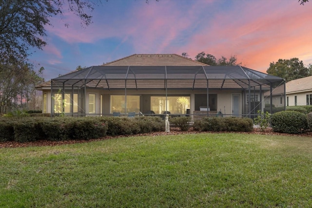 back house at dusk featuring glass enclosure and a lawn