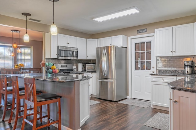 kitchen featuring dark wood-type flooring, appliances with stainless steel finishes, white cabinets, decorative light fixtures, and dark stone counters