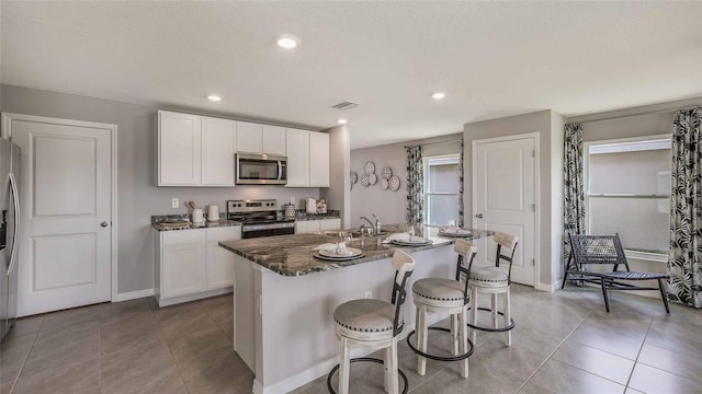 kitchen with appliances with stainless steel finishes, white cabinetry, dark stone countertops, sink, and a center island with sink