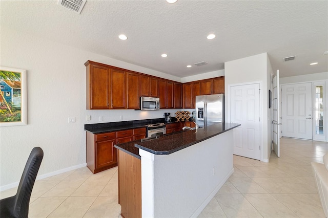 kitchen featuring a kitchen island with sink, light tile patterned flooring, dark stone countertops, and appliances with stainless steel finishes