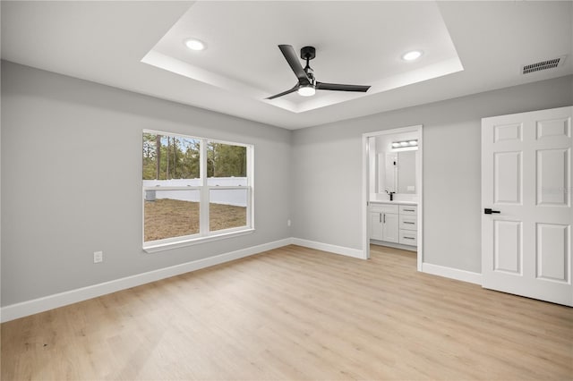 unfurnished bedroom featuring ensuite bathroom, light hardwood / wood-style floors, ceiling fan, and a tray ceiling