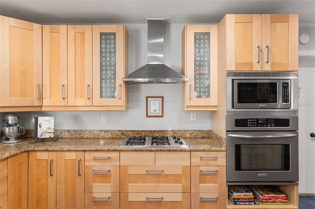 kitchen featuring appliances with stainless steel finishes, light brown cabinetry, light stone countertops, and wall chimney range hood