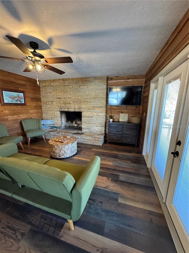 living room featuring dark wood-type flooring, a stone fireplace, wood walls, a textured ceiling, and ceiling fan