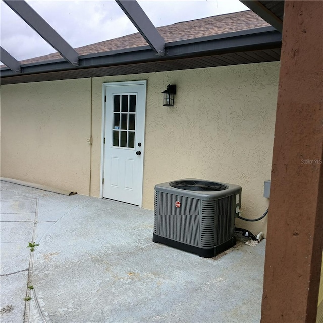 view of exterior entry featuring a patio, roof with shingles, stucco siding, and central air condition unit