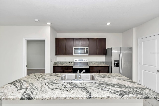 kitchen with sink, dark brown cabinets, stainless steel appliances, light stone counters, and a center island with sink