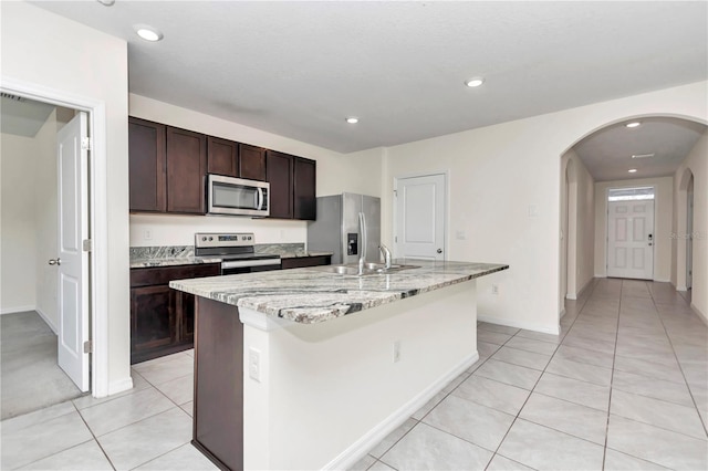 kitchen with dark brown cabinetry, sink, a center island with sink, appliances with stainless steel finishes, and light stone countertops