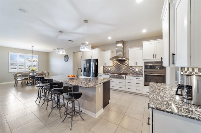 kitchen with wall chimney range hood, dark stone counters, an island with sink, stainless steel appliances, and white cabinets