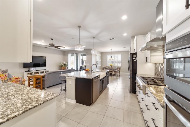 kitchen featuring pendant lighting, wall chimney range hood, white cabinetry, tasteful backsplash, and a center island with sink