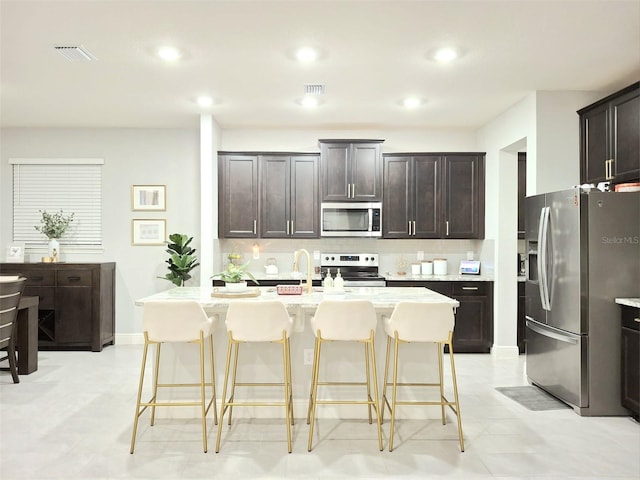 kitchen featuring a breakfast bar area, backsplash, dark brown cabinets, stainless steel appliances, and an island with sink