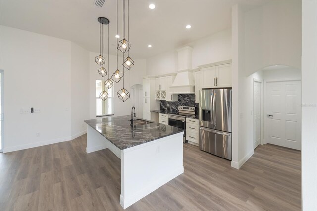 kitchen featuring custom exhaust hood, white cabinetry, decorative light fixtures, appliances with stainless steel finishes, and a kitchen island with sink