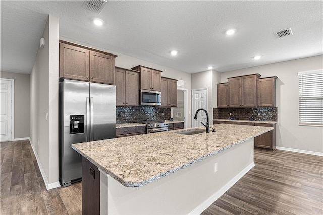 kitchen with appliances with stainless steel finishes, tasteful backsplash, sink, dark wood-type flooring, and a center island with sink