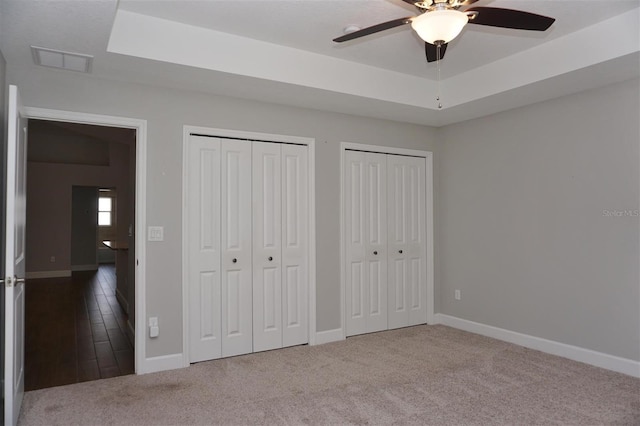 unfurnished bedroom featuring multiple closets, carpet flooring, a tray ceiling, and ceiling fan