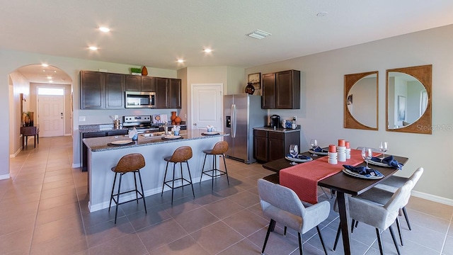 kitchen with a kitchen bar, dark brown cabinets, an island with sink, stainless steel appliances, and tile patterned flooring