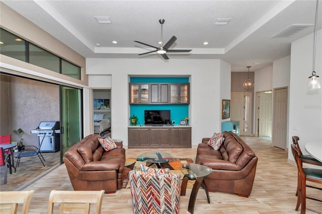 living room with a raised ceiling, ceiling fan, a textured ceiling, and light wood-type flooring