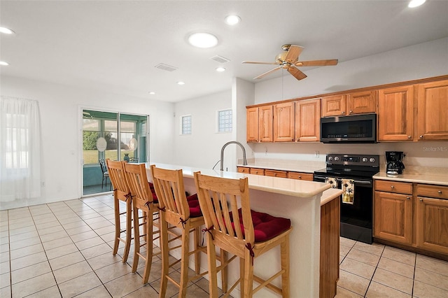 kitchen with light tile patterned flooring, a kitchen bar, an island with sink, ceiling fan, and black range with electric stovetop