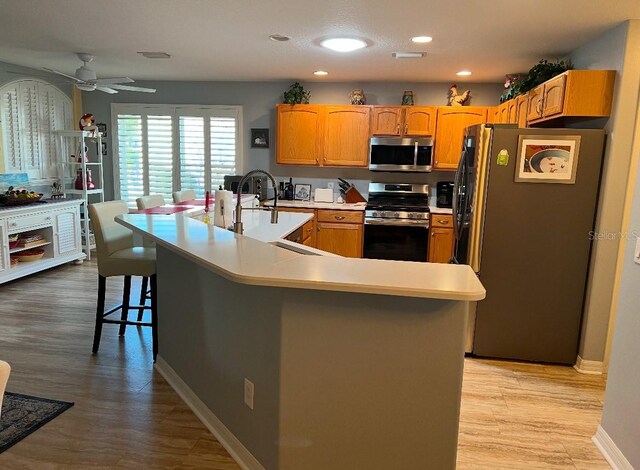 kitchen with ceiling fan, stainless steel appliances, an island with sink, a kitchen bar, and light wood-type flooring