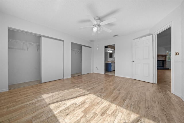 unfurnished bedroom featuring light wood-style flooring, ceiling fan, multiple closets, a brick fireplace, and connected bathroom