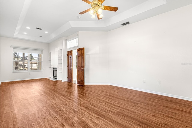 unfurnished living room with a tray ceiling, wood-type flooring, built in shelves, and ceiling fan