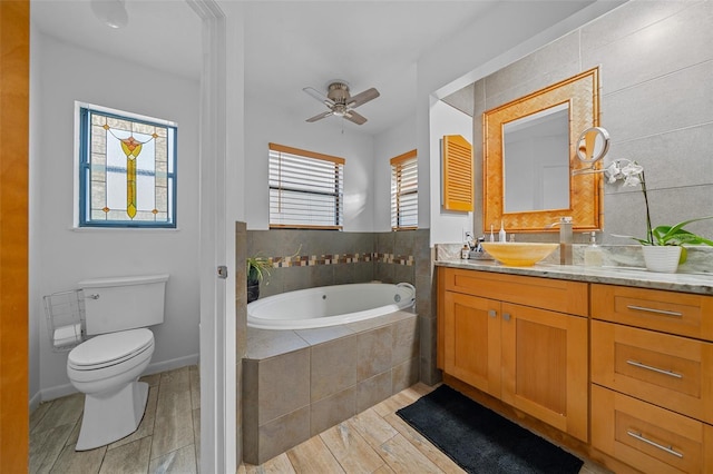 bathroom with tiled tub, vanity, ceiling fan, and toilet