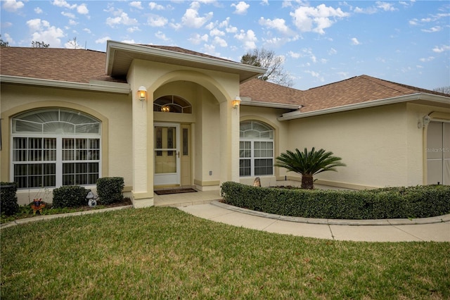 entrance to property with stucco siding, a lawn, and roof with shingles