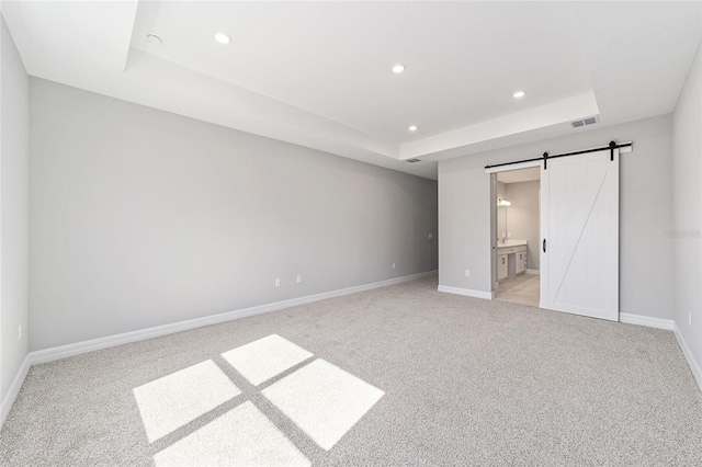 unfurnished bedroom featuring a tray ceiling, light carpet, ensuite bathroom, and a barn door