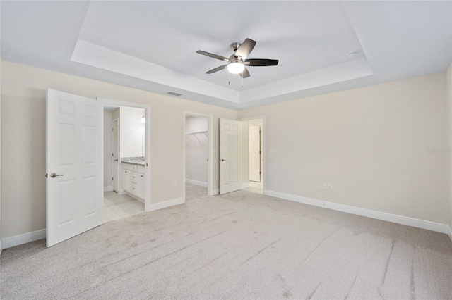 unfurnished bedroom featuring a walk in closet, ensuite bathroom, ceiling fan, a raised ceiling, and light carpet