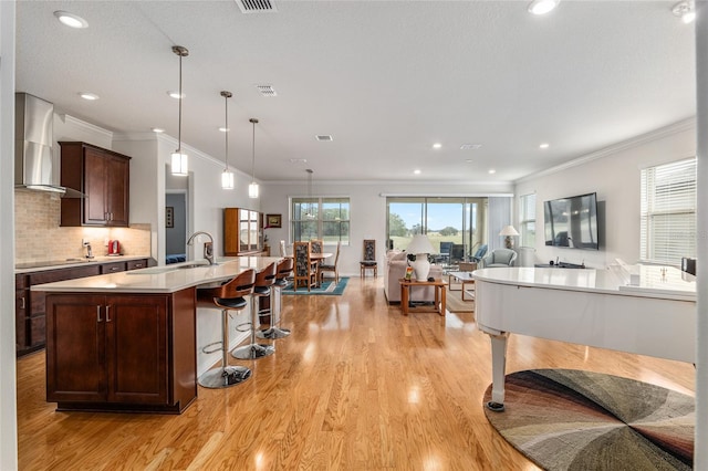 kitchen with light hardwood / wood-style flooring, a kitchen island with sink, hanging light fixtures, dark brown cabinetry, and wall chimney exhaust hood