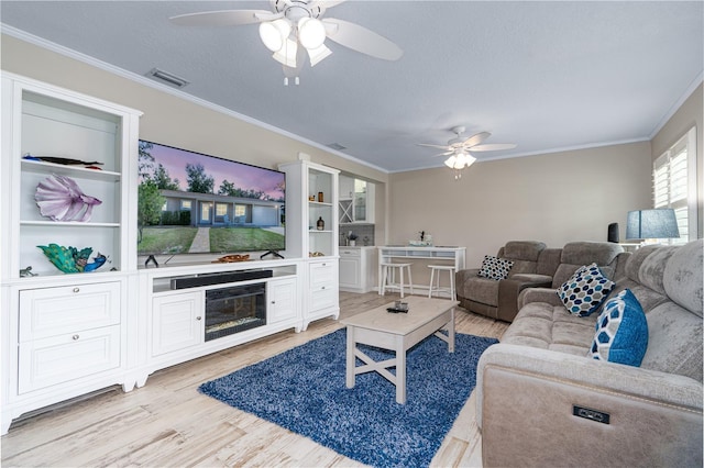 living area with a ceiling fan, visible vents, ornamental molding, light wood-style floors, and a glass covered fireplace