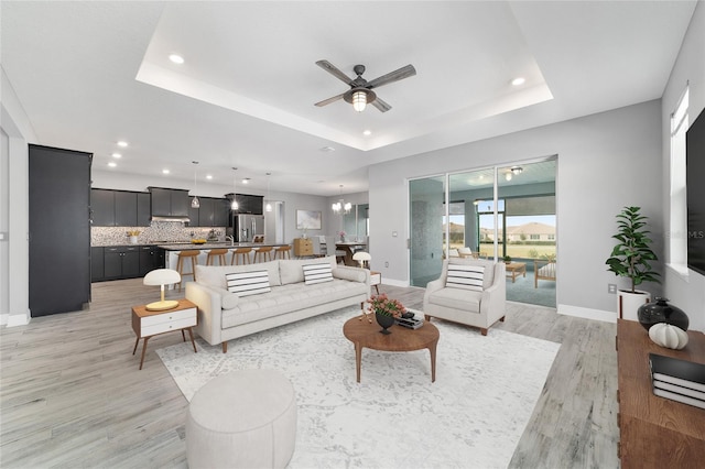 living room with a tray ceiling, light wood finished floors, recessed lighting, baseboards, and ceiling fan with notable chandelier