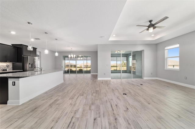 unfurnished living room with light wood-style flooring, a tray ceiling, baseboards, and ceiling fan with notable chandelier